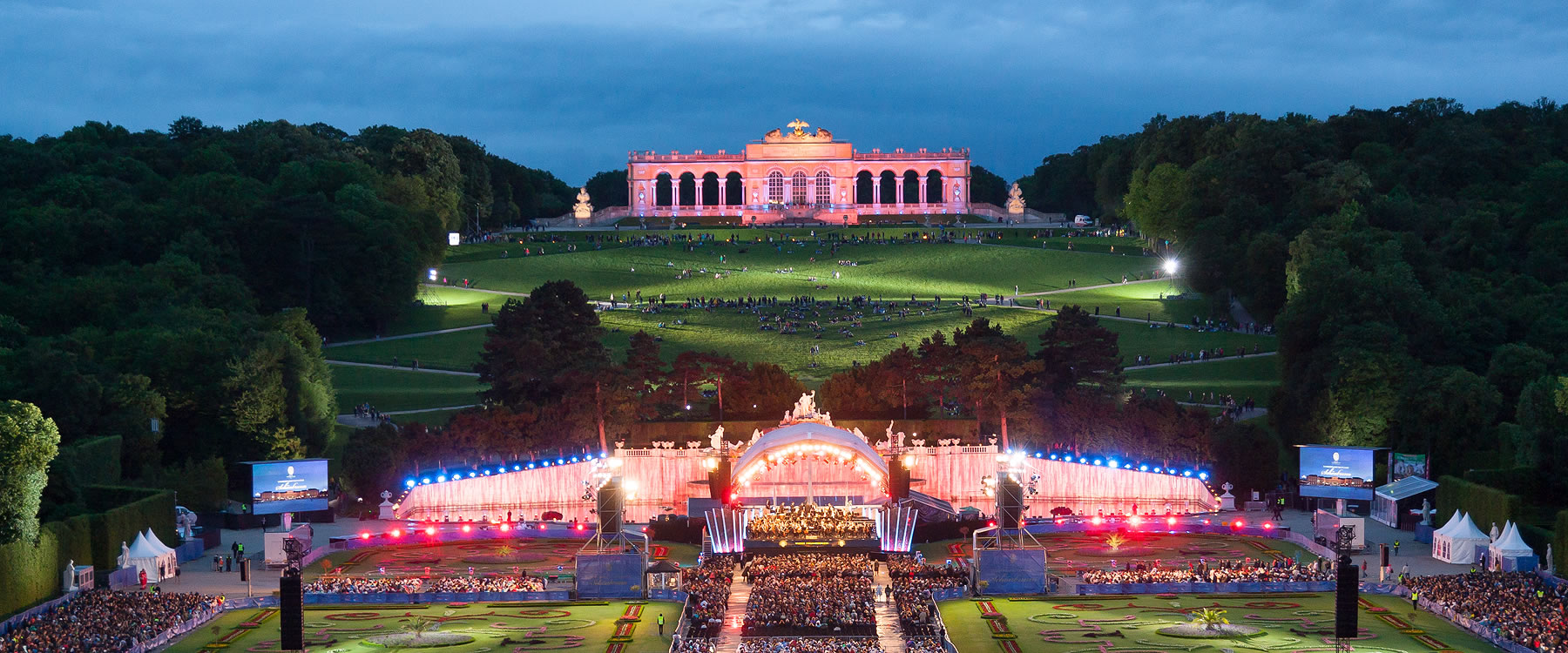 outdoor evening concert with orchestra in mid-ground and large palace in background