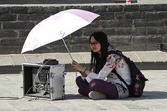 Crew member seated on the ground holding an umbrella over equipment